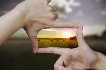 Fingers framing clear portion of image of green field with setting sun